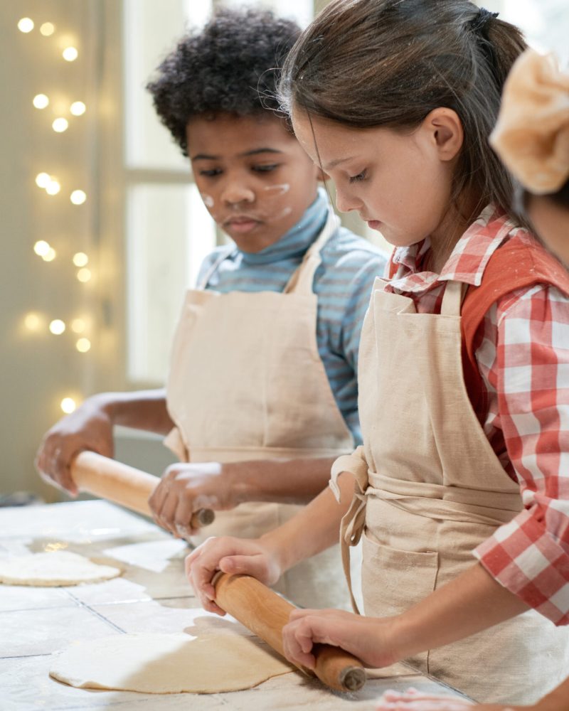 Group of children in aprons rolling the dough with rolling pin at the table during cooking lesson with the chef